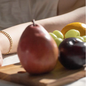 A hand wearing a gold-colored bracelet rests next to a wooden board with apples and grapes on it.