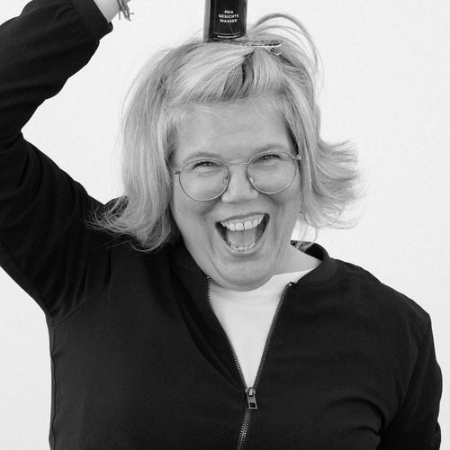 A woman with a cheerful expression stands smiling against a neutral background. Balancing on her head is a brown glass bottle with