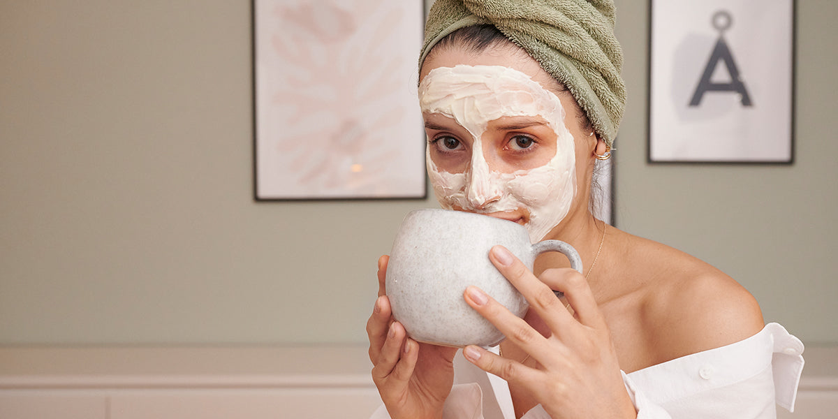 A woman drinking from a teacup with a face mask to face and Towel wrapped around her head.