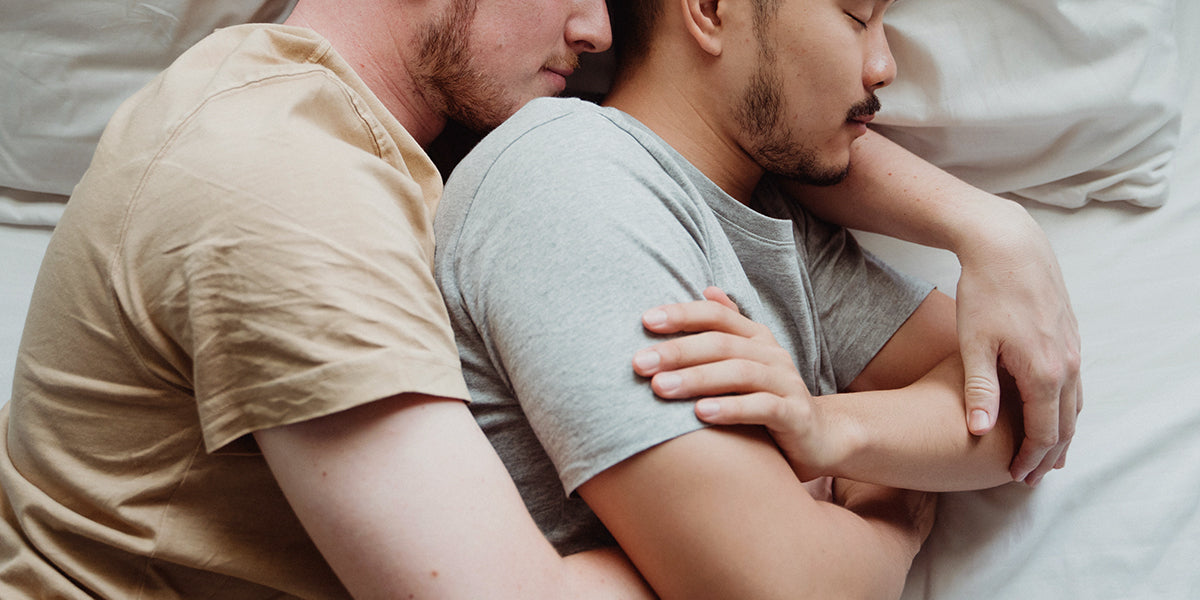 Two men lying asleep in bed, holding each other.
