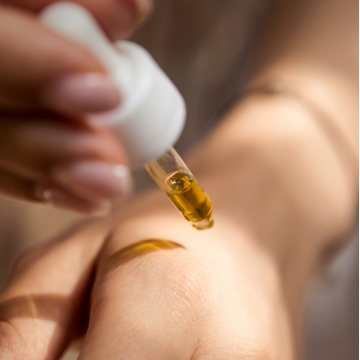 A hand with a delicate skin tone, onto which a drops golden oil drips from a pipette.
