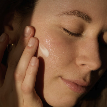 A woman is applying a light layer of cream to her cheek; her eyes are closed. Her natural skin texture is visible.