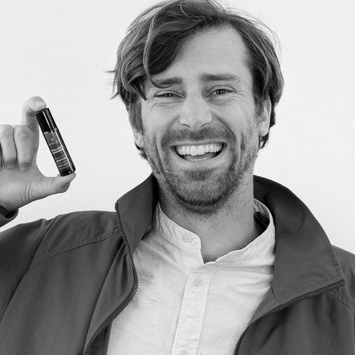 A man is smiling as he holds a product in a small tube; he is wearing a light-colored blouse and a jacket.
