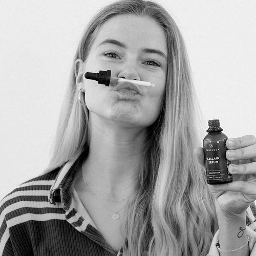 A woman is holding an amber glass bottle with a dropper and smiling. She is wearing a striped blouse and has long,