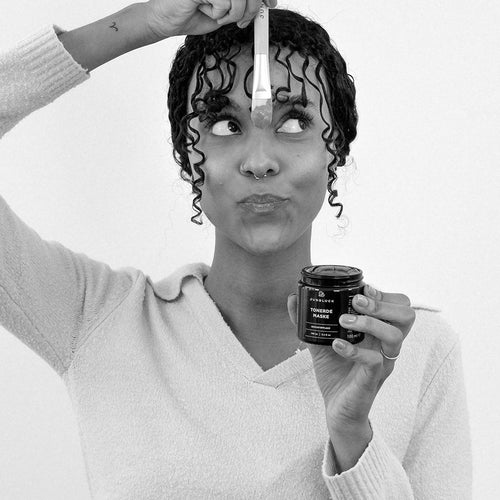 A woman with curly hair is holding a paintbrush and a brown glass container. She is wearing a light-colored sweater and is looking