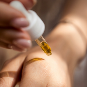 A hand with a delicate skin tone, onto which a drops golden oil drips from a pipette.