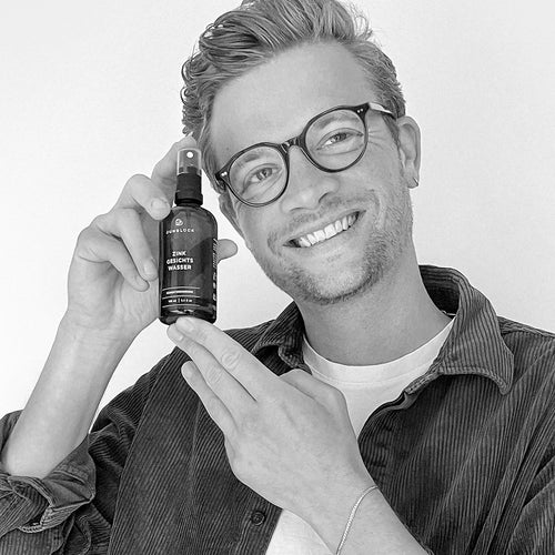 A man is smiling as he holds a brown glass bottle containing the product "Matze" while looking into the camera.