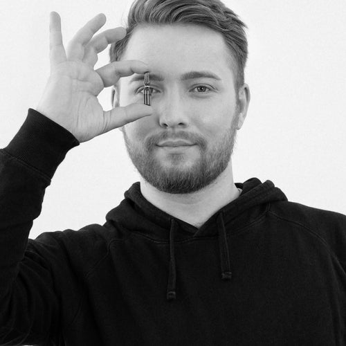 A young man holds a small amber glass bottle up to his eye and looks into the camera. He is wearing a black