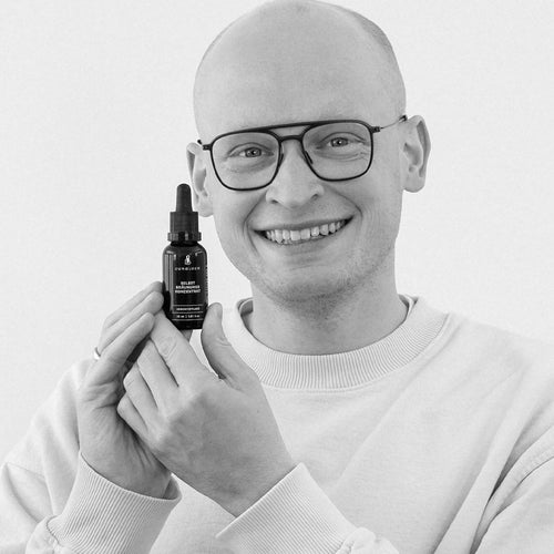A man is holding a brown glass bottle containing the product "Benedikt" and smiling at the camera.
