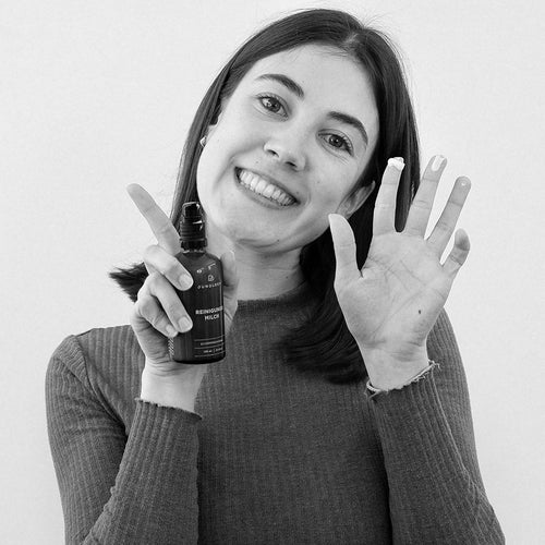A person is holding a product in an amber glass bottle with both hands and smiling. The background is bright and neutral.